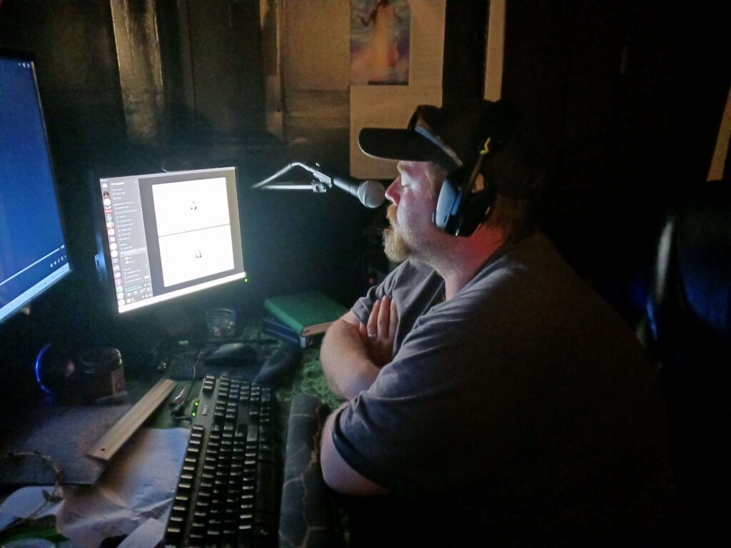 Candid photo of Damon Bryant, a white male with dark shirt, dark ball cap, red-brown goatee, wearing headphones at his desk speaking into a microphone lit by the light of 2 computer monitors.
